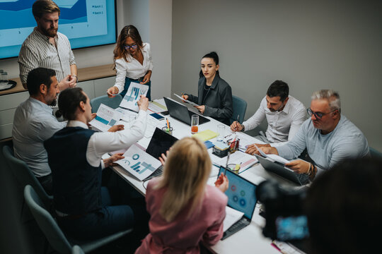 A diverse group of colleagues gathers for a data-driven meeting, reviewing charts, laptops, and documents. They discuss strategies, share ideas, and collaborate across a modern office setting. - Powered by Adobe