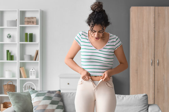 Upset African-American woman trying to button tight pants at home. Diet concept