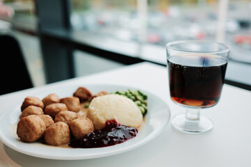 Plate of Swedish meatballs with mashed potatoes, green peas, and gravy served beside a glass of soda on a white table by the window