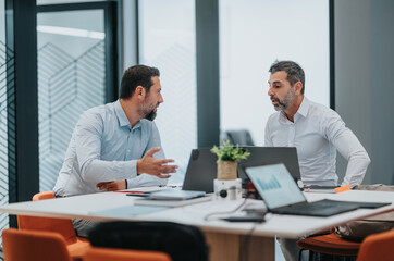 Obraz premium Two colleagues engage in a focused discussion at a contemporary office, with laptops, charts, and a small plant on the table. The scene conveys collaboration, planning, and professional teamwork.