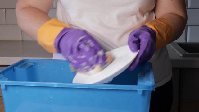 Close-up of a woman scrubbing a plate with a sponge inside a blue basin, wearing purple and yellow gloves due to a non-working faucet