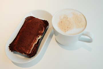 Classic tiramisu topped with cocoa powder served beside a foamy cappuccino on a white table, minimalist cafe setting. The concept of breakfast in a cafe