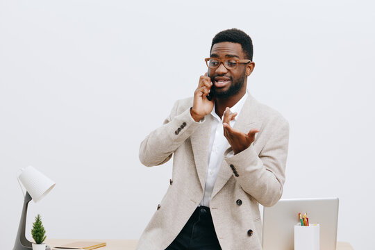 Confident young African-American man in beige blazer and glasses talking on smartphone while gesturing with hand in bright office interior, professional business lifestyle concept - Powered by Adobe
