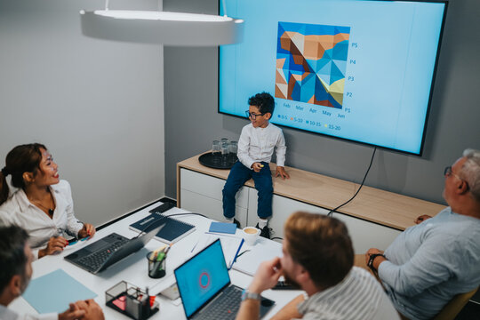 A diverse group gathers around a table as a young boy explains a colorful graph projected on a screen, illustrating collaboration, learning, and teamwork in a contemporary office setting. - Powered by Adobe