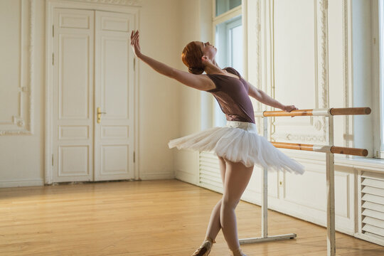 Young ballerina in tutu skirt stretching practicing ballet positions dancing near ballet barre in dance class. Young classical dancer woman in dance studio. Ballet class training Ballet school concept