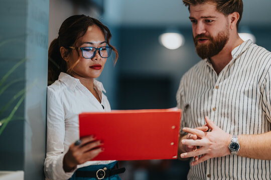 Two professionals examine a red folder in a modern office, sharing ideas and staying focused on the task. A scene of teamwork, communication, and professional collaboration between a man and a woman.