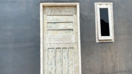 An old off-white wooden door on a gray wall with a small window beside it