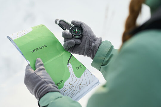 Caucasian young adult woman holding map and compass, navigating outdoors with gloved hands, focusing on route through forest area, partial view showing braided hair and outdoor clothing