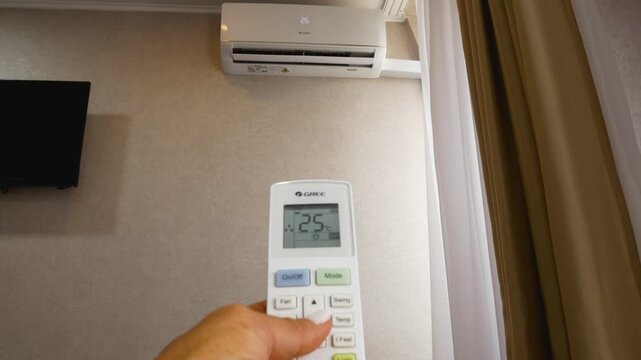 Remote control in hand opposite the air conditioner, which hangs on the wall. The display of the remote control shows how the temperature in the room is lowered, the air conditioner is set with the