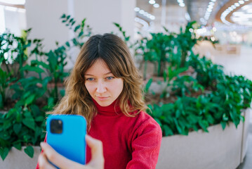 Young woman in a red turtleneck, focusing on her smartphone screen while taking a selfie for social media, surrounded by indoor plants in a contemporary shopping mall
