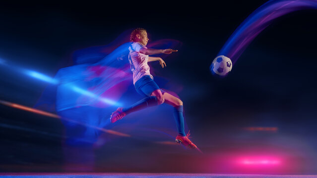 Female soccer player jumping for kick under neon motion lights at night stadium. Concept of fitness promotion, sport branding visuals, advertising for training and active lifestyle.