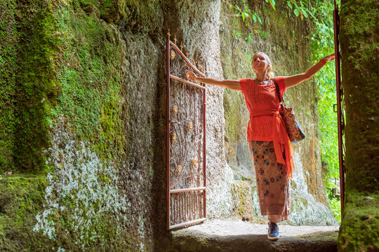 Woman in traditional sarong enters ancient temple gate in Gunung Kawi, Bali, surrounded by mossy rock and tropical greenery — symbolizing peaceful exploration of cultural and sacred places. - Powered by Adobe