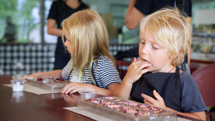 Two kids enjoy chocolate making class in Bali. Little boy happily tastes his sweet creation while girl focuses on decorating. A fun and flavorful family experience during travel