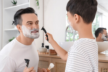 Happy father and his little son holding razors with shaving foam in bathroom