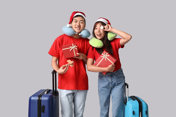 Beautiful young Asian couple in Santa hats with gift boxes, wooden airplane and suitcases on grey...