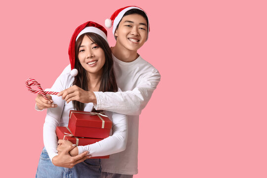 Beautiful young Asian couple in Santa hats with gift boxes and candy canes on pink background. Christmas celebration - Powered by Adobe