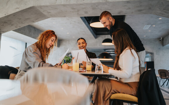 A group of four friends or colleagues gather around a table in a modern cafe, reviewing documents and sharing ideas while smiling and enjoying orange juice. - Powered by Adobe