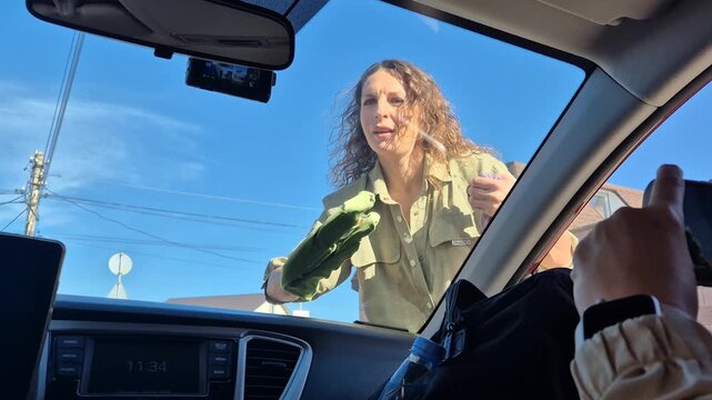 Woman cleaning windshield of vehicle on sunny street, providing car washing services for driver.