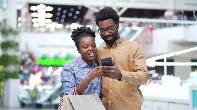 Happy African American family couple shoppers using mobile phone application standing in shopping center mall. Smiling man woman buyers use online app store for discount, sale promotions on smartphone