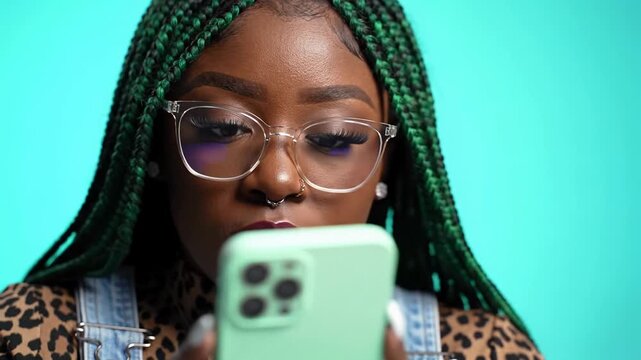 Young Black woman with vibrant green box braids and chic glasses, in a leopard print top, intently gazing at a glowing smartphone. Plain teal background, slow push-in. Digital immersion