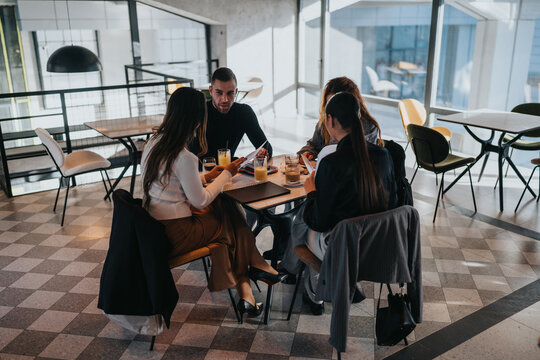 A diverse group of friends and colleagues sit around a table in a bright cafe, discussing work with notebooks and drinks. Casual yet focused business meeting in a contemporary setting.