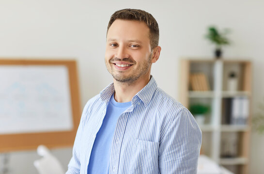 Portrait of cheerful smiling bearded man in casual wear standing in relaxed posture on workplace and looking at camera. Positive caucasian employee in light blue shirt in modern office or classroom. - Powered by Adobe