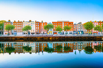 A colorful row of historic buildings along the River Liffey in Dublin is beautifully reflected in the calm water under a clear blue sky.