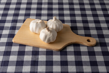 Top view of three whole garlic bulbs resting on a light-colored wooden cutting board on a navy blue and white plaid tablecloth. Ideal for cooking, ingredients, and food preparation concepts.