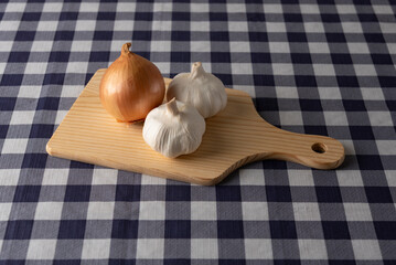 Top view of one onion and two garlic bulbs on a light-colored wooden cutting board on a navy blue plaid tablecloth. Ideal for cooking, ingredients, and food preparation concepts.