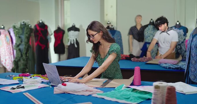 Indian young adult gen z female lady wear tape around neck learn clothe craft skill indoor class room. girl use laptop look screen typing hold pencil do fabric pattern on paper new day college place