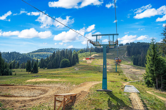 A vibrant summer landscape featuring a chairlift ascent over grassy slopes and forests under a bright blue sky with white clouds.
