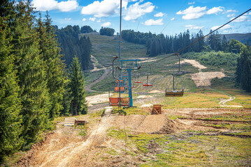 A vibrant summer landscape featuring a chairlift ascent over grassy slopes and forests under a bright blue sky with white clouds.