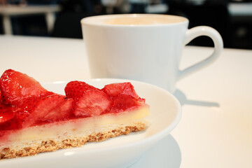 Close-up of glazed strawberry tart with a cup of cappuccino in the background on a white table in a cozy cafe