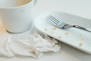Empty cup with coffee foam, white plate with crumbs, fork, and crumpled napkin after a meal