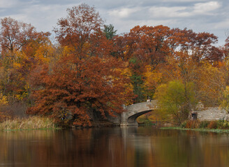 Beautiful fall colors with orange and yellow trees surrounding a serene pond with a stone bridge in Verona Park New Jersey during autumn season