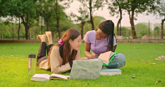 Indian happy young adult female talk use laptop look screen check online work task share idea plan gen z girl lying on grass do study help read new exam course book enjoy spend day time park place