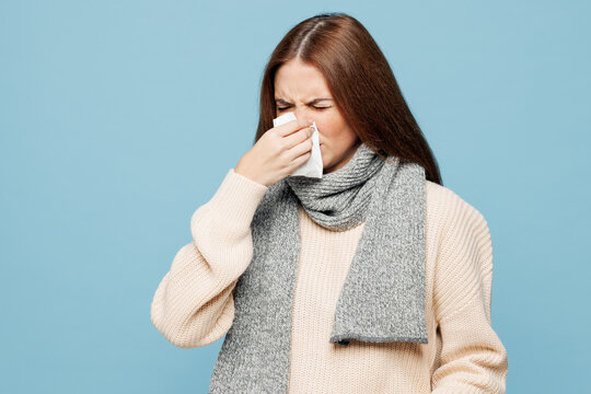 Young woman wear pink sweater scarf hold in hand paper tissue blowing wiping nose isolated on plain blue background studio portrait. Healthy lifestyle ill sick disease treatment cold season concept.