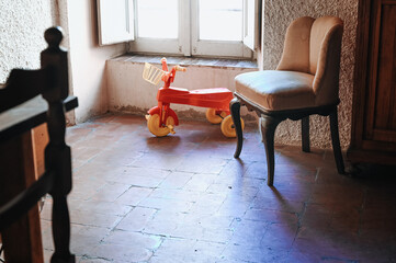 In a dark room illuminated by soft light from the window stands an old vintage red children’s bicycle. An atmospheric and nostalgic scene filled with warmth and silence.
