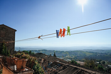 Four colorful clothespins hanging on a clothesline on a terrace, with a beautiful view of the Italian countryside, mountains, and fields in the background under a clear sky.
