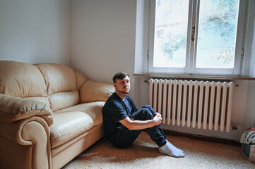 A man sitting on a beige sofa at home, holding a hat in his hands, looking thoughtful. Warm interior lighting, quiet atmosphere, comfort, and introspection.