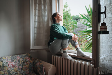 Relaxed woman sits on a windowsill surrounded by sunlight and green plants, enjoying peace and quiet morning moments at home.
