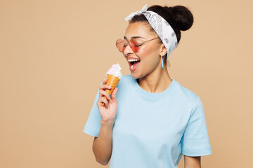 Young smiling happy woman of African American ethnicity wears blue t-shirt casual clothes hold in hand eat vanilla ice-cream isolated on plain pastel light beige background studio. Lifestyle concept.