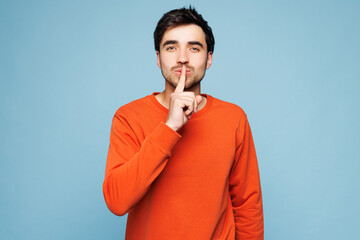 Young Caucasian man he wear orange sweatshirt casual clothes say hush be quiet with finger on lips...