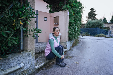 Young woman in a purple dress sits on the corner of a quiet Italian street, surrounded by warm tones and greenery, enjoying a peaceful evening.
