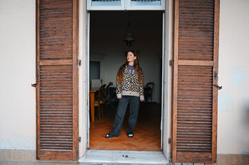Young woman stands in a doorway, dressed casually in a patterned shirt and jeans, illuminated by soft natural light from the outside.
