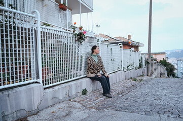 Calm woman in a warm jacket sits on a street bench near a white metal fence, enjoying a quiet moment in a small Italian neighborhood.
