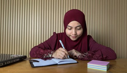 Young woman wearing hijab writing in notebook at desk
