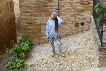 Stylish young woman wearing a blue hoodie and checkered pants stands on a cobblestone street, enjoying the quiet charm of an Italian old town.
