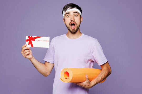 Young shocked strong fitness trainer man sportsman he wearing t-shirt train in gym hold yoga mat gift coupon voucher card for store isolated on plain purple background. Workout sport fit abs concept.