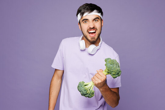 Young vegan fitness trainer instructor sporty man sportsman he wearing headphones t-shirt train in home gym hold broccoli pov dumbbells isolated on plain purple background. Workout sport abs concept.